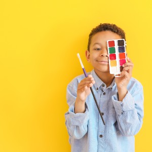 Black boy artist with watercolors and paintbrush in front of a yellow background