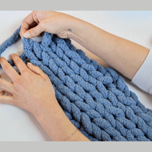 A blue knitted scarf on a grey background. Two hands are working on it.