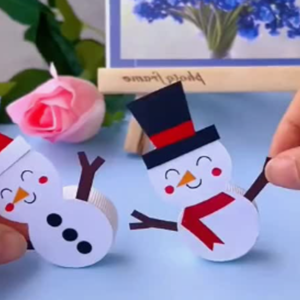 A paper and plastic snowman sits on a white background. It is tipped to one side as though waving